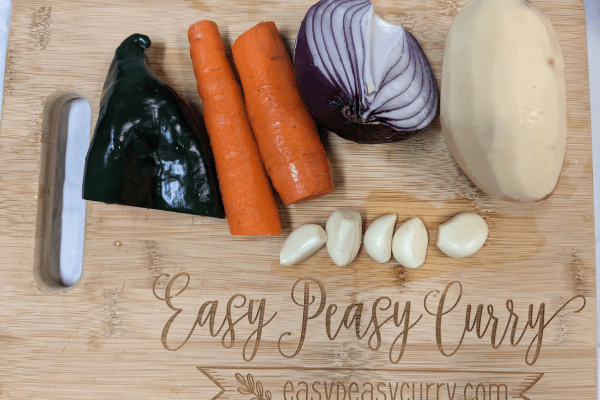 Garlic cloves, half a red onion, potato, carrot and poblano pepper on a cutting board